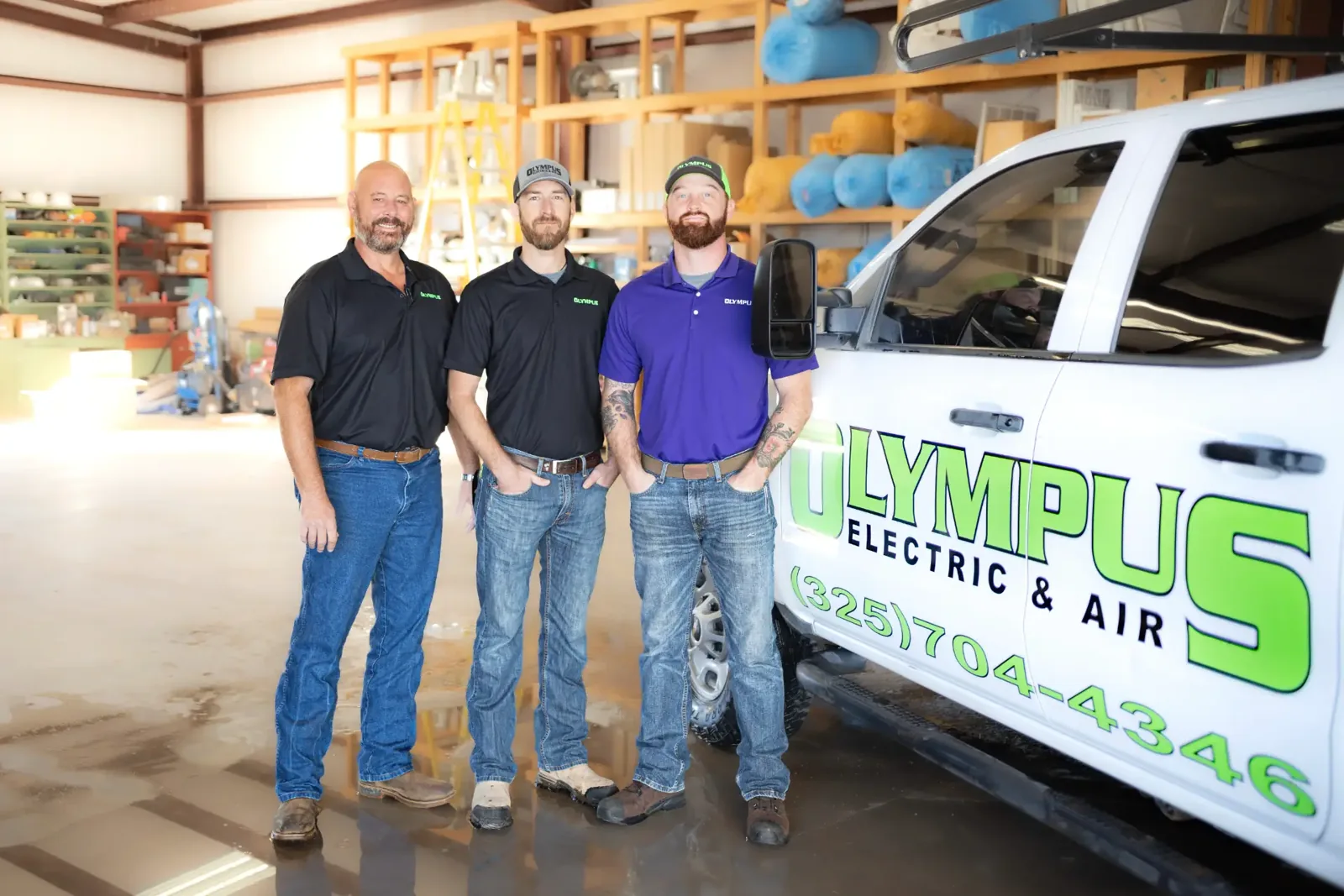 Three Olympus staff beside a branded Olympus Electric & Air truck in the shop